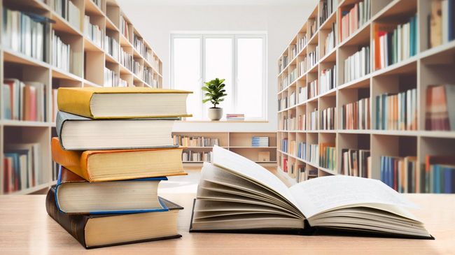 A stack of books and an open book sit on a wooden table in a bright library with bookshelves and a window in the background.