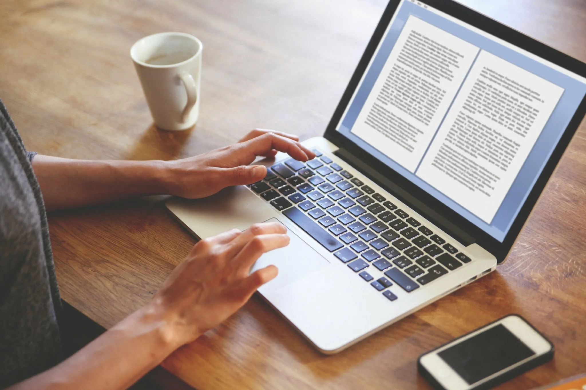 Person typing on a laptop with a document on screen, coffee cup and phone on wooden table beside them.